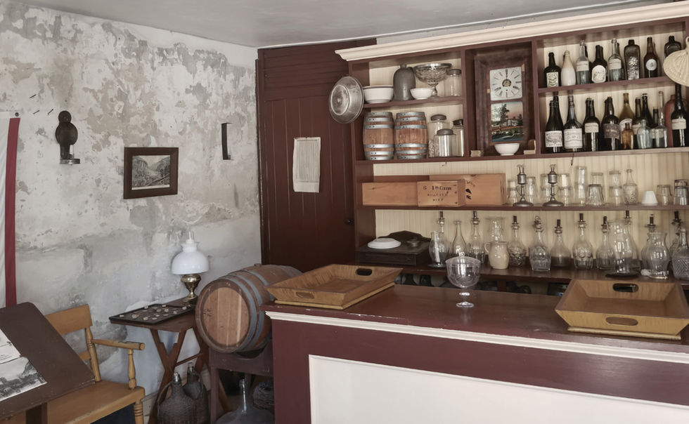 Interior of a historic tavern in Harpers Ferry with wooden shelves filled with bottles, jars, and pots; a countertop with a goblet and cutting board, a table with an oil lamp, aged walls, and vintage furnishings—evoking frontier hospitality and domestic rhythm.