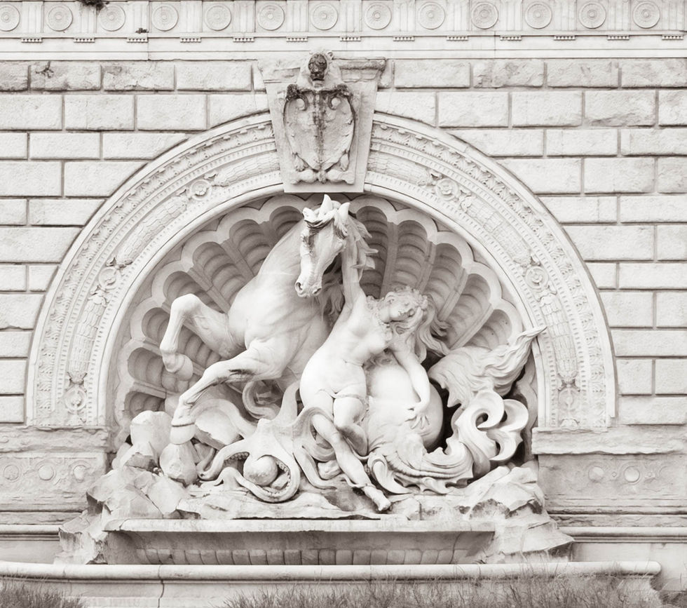 Relief sculpture of a human figure interacting with a rearing horse, framed by swirling marine motifs and set in a scallop-shell niche on a brick wall; a crest with shield decorates the top, suggesting mythological or civic symbolism.