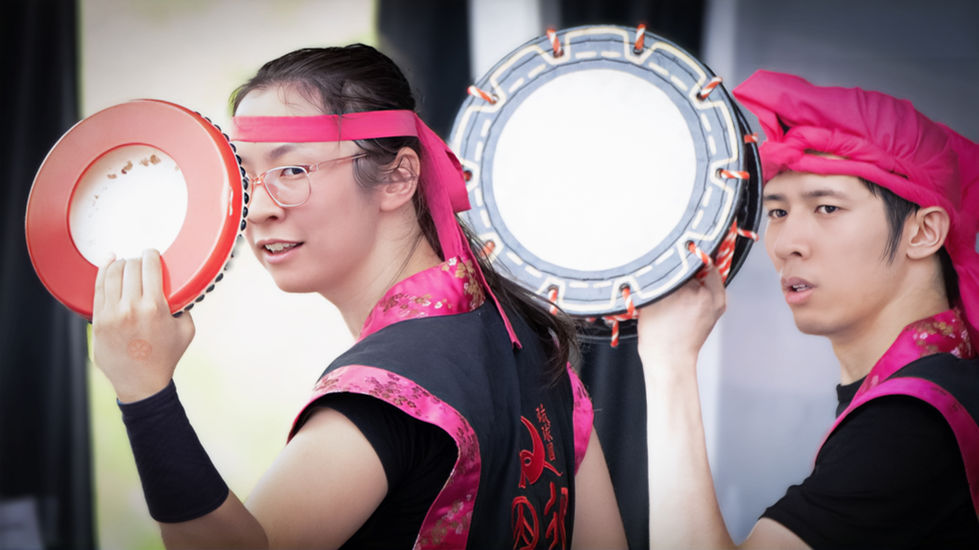 Two performers at Sakura Festival wearing black outfits with pink trim, each holding a handheld drum; one wears a pink headband and glasses, the other a pink wrap, in a moment of cultural and musical expression.