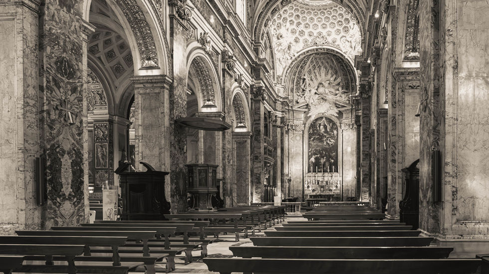 Black-and-white interior of a cathedral featuring vaulted ceilings, decorated columns, frescoed arches, a central aisle with pews, and an ornate altar with religious artwork—evoking sacred atmosphere and architectural grandeur.