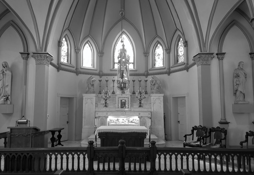 Interior of a Gothic-style chapel in Harpers Ferry with pointed arches, vaulted ceiling, crucifix, stained glass windows, altar marked “ALMS,” and surrounding pews and chairs—evoking sacred architecture and historical reverence