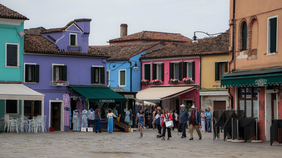 Colorful street scene in Burano, Italy, with brightly painted buildings in purple, blue, pink, yellow, and orange; tiled roofs, shuttered windows, and awnings frame a lively crowd in a market or tourist area.