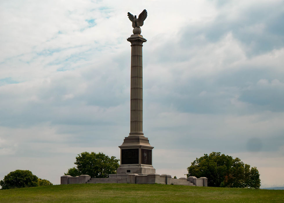 Tall column monument at Antietam Battlefield topped with a spread-winged eagle; stone base includes inscriptions, surrounded by trees and cloudy sky—symbolizing vigilance and Civil War remembrance.