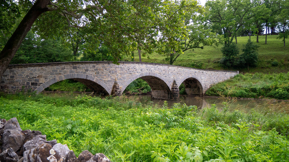 Historic stone bridge with four arches spanning a calm creek, surrounded by grass, trees, and rural landscape—known as Burnside Bridge at Antietam Battlefield, symbolizing tactical struggle and historical significance