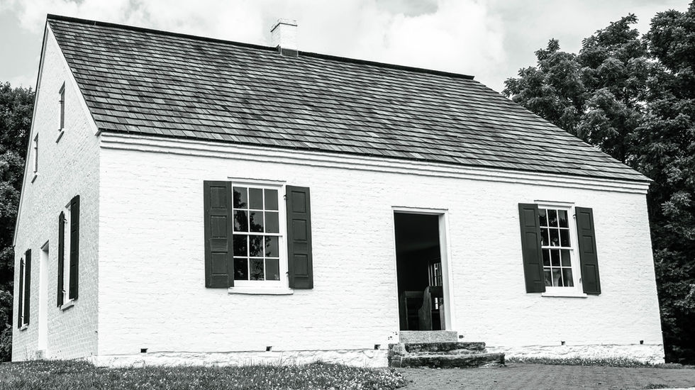 White-painted brick building with steep shingled roof, three shuttered windows, and central doorway with steps; rural setting with trees in the background—known as Dunker Church, a historic site at Antietam Battlefield