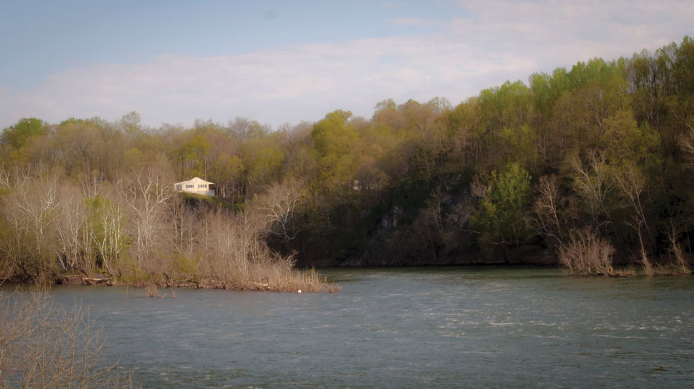 Lakeside scene along the C&O Canal with calm water in the foreground, early spring trees with mixed foliage, and a small cabin with a light roof partially visible in the background under a clear sky.