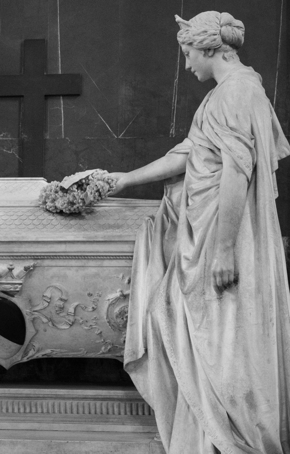 Woman placing flowers on marble tomb, with cross and sculptures in background.