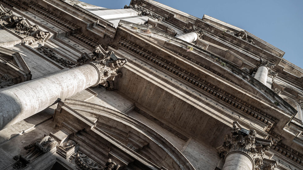 Upward view of a classical building with ornate columns, decorative cornices, and aged stonework; dramatic angle highlights architectural detail and historical craftsmanship.