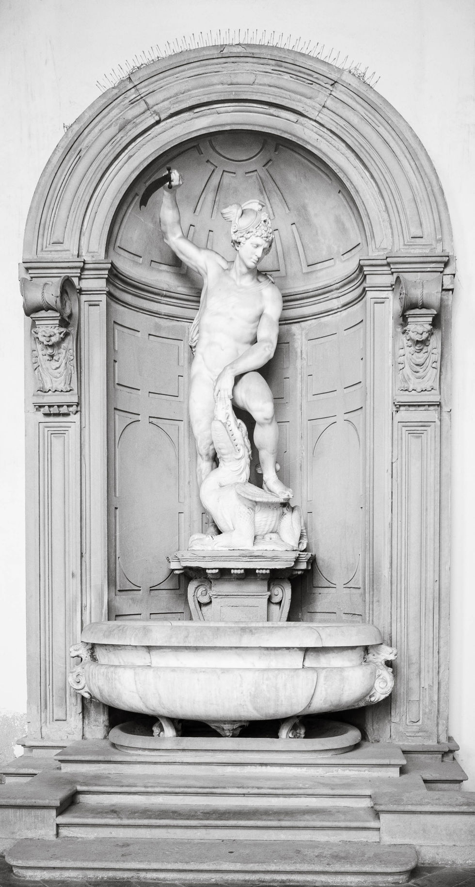 Grayscale image of a classical stone fountain featuring a robed figure in a niche, one arm raised and the other holding a circular object; architectural framing includes columns and arch, with a basin below.