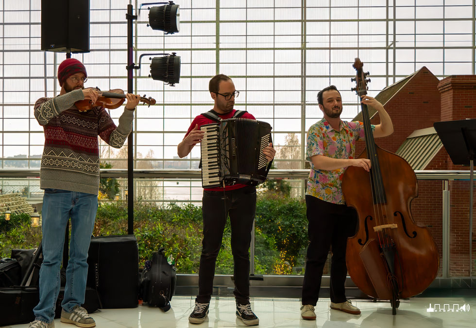 Three musicians performing indoors at MAGFest in front of a large grid-patterned window with greenery outside—one playing violin in a red beanie and patterned sweater, one playing accordion in a red and black shirt, and one playing double bass in a floral shirt, with stage lights positioned behind them