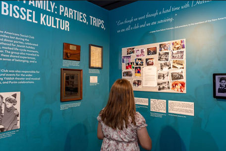 A child stands with her back to the camera, looking over a museum exhibition made up of wall text, artifacts, and photos