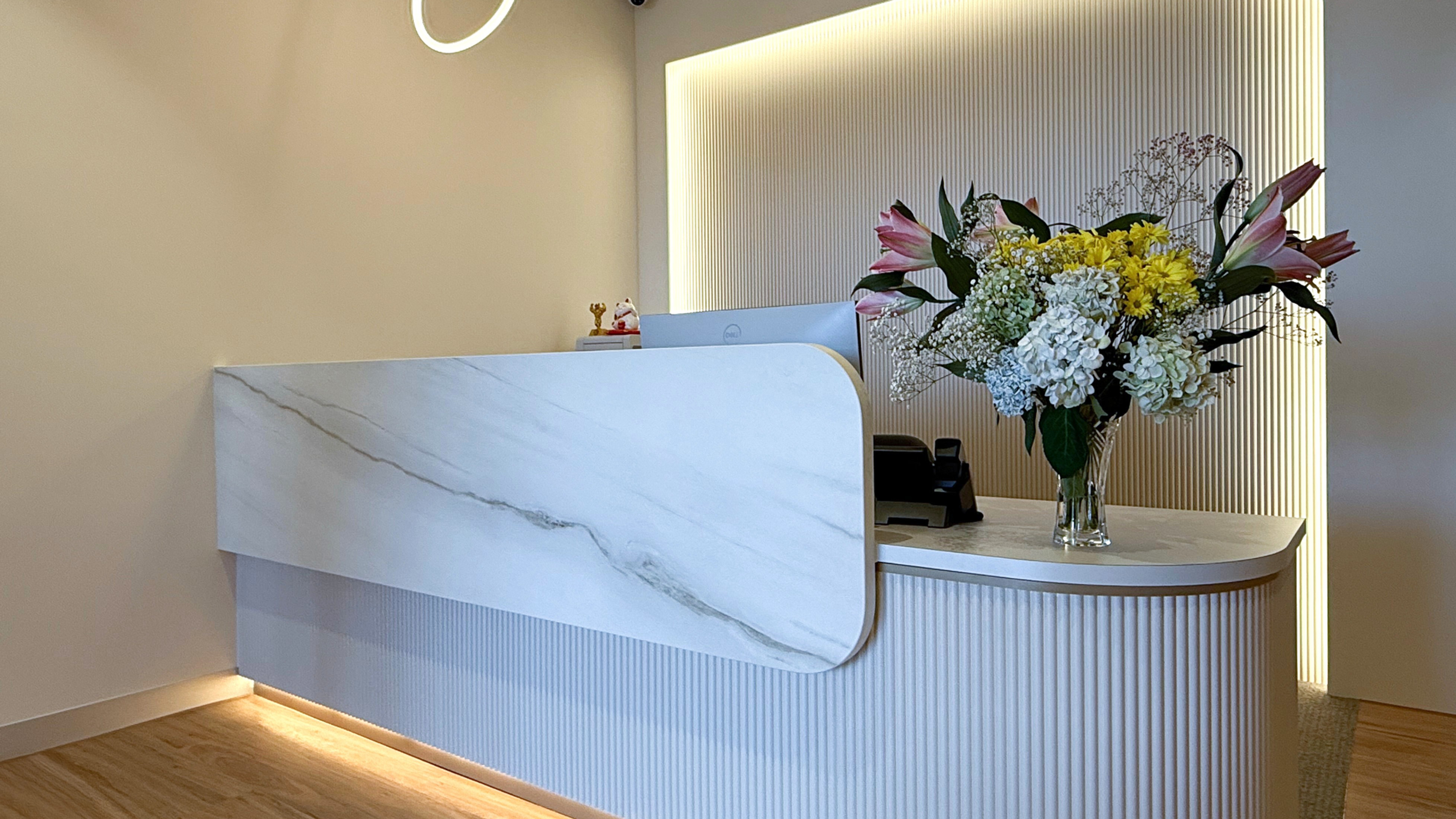 Modern kitchen reception desk featuring white marble counter with a curved fluted wooden panel base, softly illuminated by concealed LED strip lighting, with a vibrant floral arrangement in a glass vase.