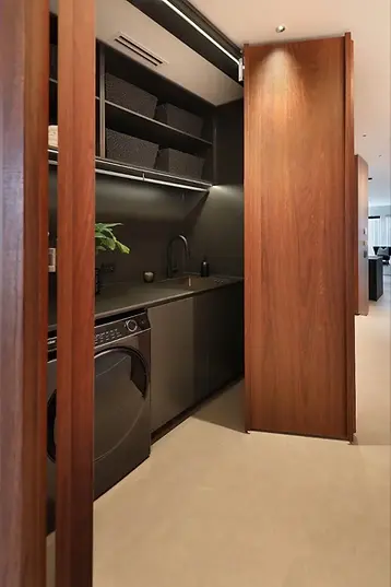 Concealed matte black European laundry joinery revealed behind full-height walnut timber veneer bifold door, featuring matte black cabinetry, open shelving with dark storage baskets, integrated hanging rail, matte black undermount sink with gooseneck tapware, sintered stone benchtop, and front-loader washing machine — bespoke concealed laundry joinery by Premier Form Pty Ltd, Melbourne Victoria Australia