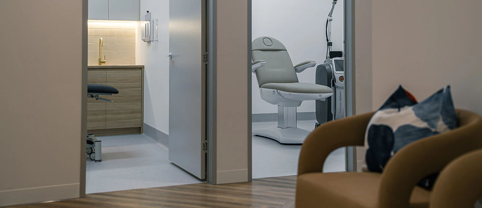 Purpose-built medical aesthetic clinic fitout showing dual treatment room view through open pivot doors — left room features custom oak timber veneer floating vanity joinery with brushed gold gooseneck tapware, white subway tile splashback with integrated warm LED strip lighting under wall-mounted tri-fold mirror, and clinical treatment bed; right room features adjustable cream upholstered medical treatment chair, professional laser skin treatment machine on articulating arm, vinyl clinical flooring, and LED panel ceiling lighting — bespoke medical aesthetic clinic joinery and fitout by Premier Form Pty Ltd, Malvern East Melbourne Victoria Australia