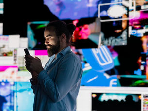 A man in a blue shirt standing in profile, looking at a smartphone against a background of multiple glowing digital screens displaying social media and video content.