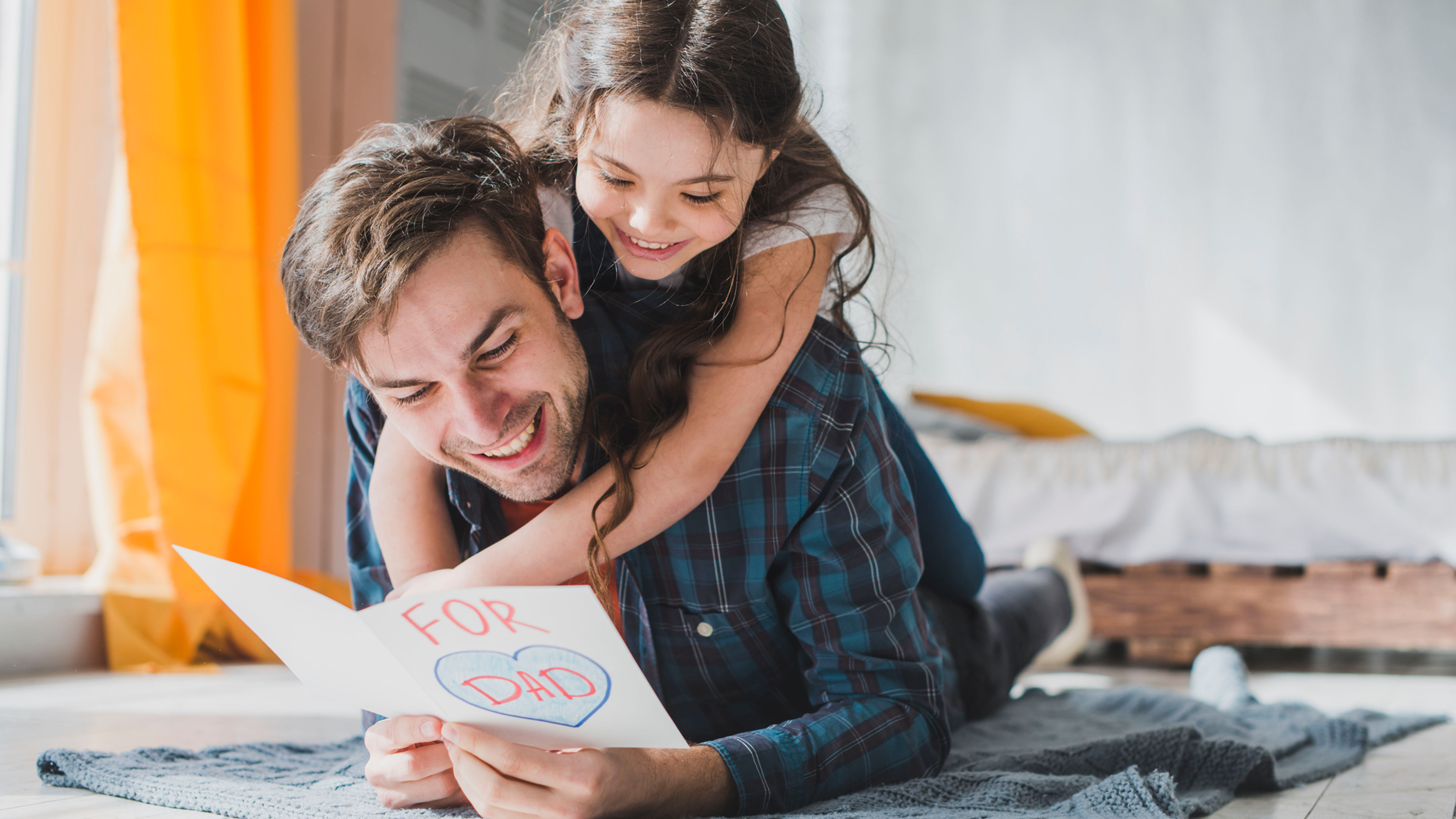 Smiling girl hugs dad as he reads a handmade card saying FOR DAD on the floor of a bright room.