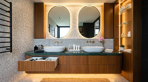 Curved floating timber veneer double vanity  with Verde green marble benchtop, integrated warm  LED underglow lighting, dual white vessel basins,  brushed brass wall-mounted tapware, backlit oval  black-framed mirrors, penny round mosaic tile  feature wall, and open timber storage tower with  glass shelves — luxury master ensuite double vanity  joinery by Premier Form Pty Ltd, Melbourne  Victoria Australia