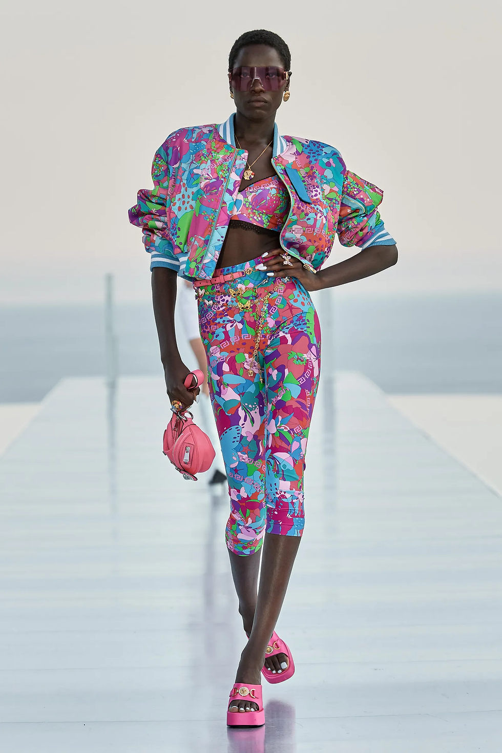 La Vacanza Dua Lipa x Versace La Vacanza multicoloured pedal pushers, bralette and jacket with bright barbie pink handbag and platform shoes.