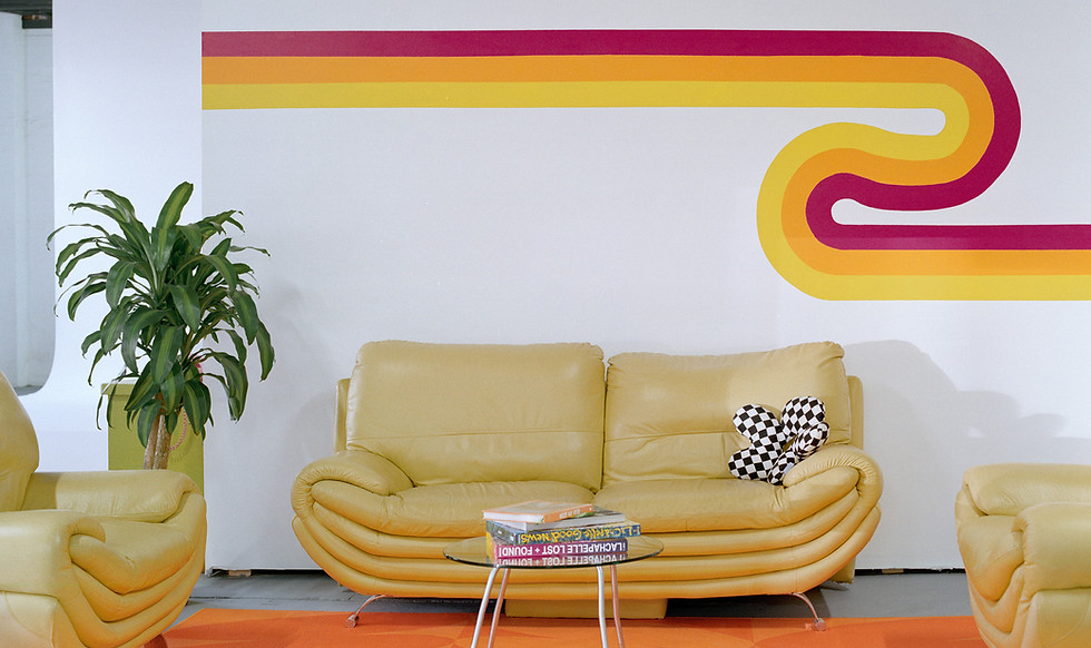A colourful loungeroom with yellow couches, an orange rug and a plant. Behind on the wall is a colourful striped retro mural.