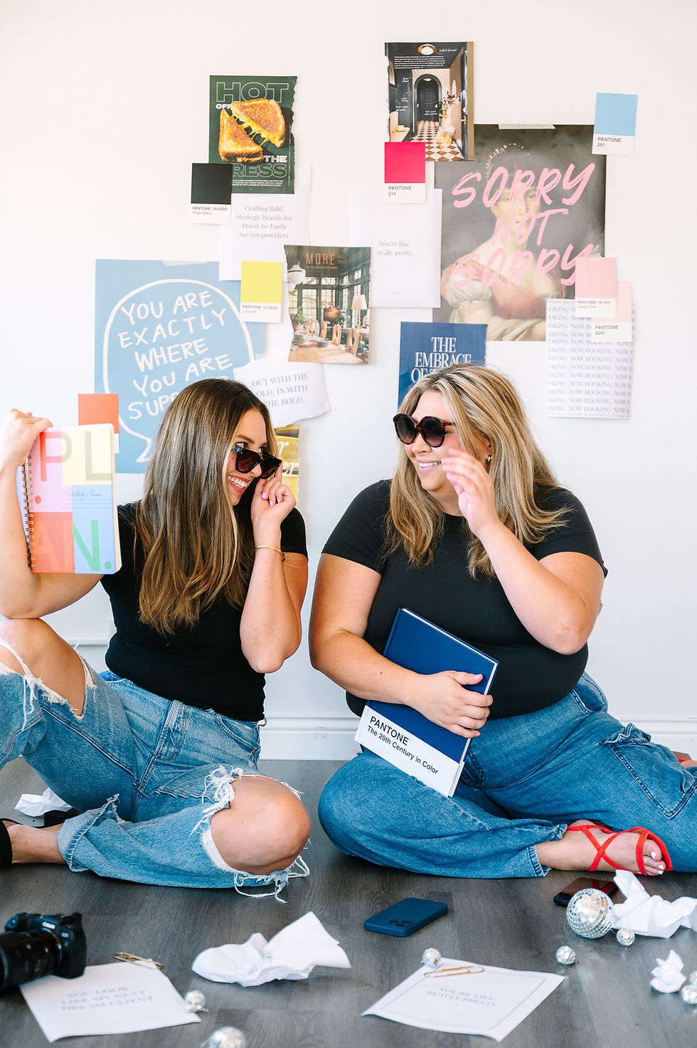 Two women in sunglasses and jeans sit on the floor, smiling. Behind them is a collage with colorful posters. Text includes "PLAN" and "PANTONE".