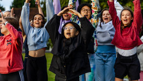 Group of children raising their arms joyfully outdoors. They wear colorful jackets and glasses. Trees in the background, vibrant mood.