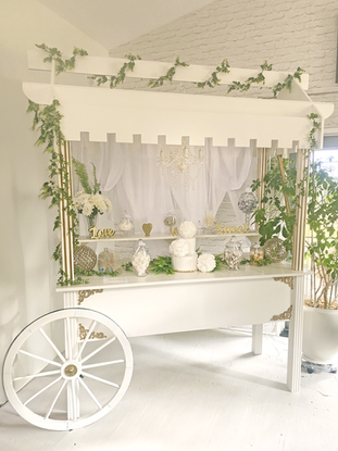 White and gold rental Treats Cart, decorated with a selection of white and gold candy in glass serving bowls, decorated with greenery.