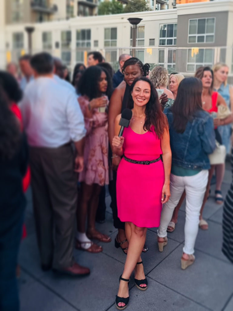 A woman in a bright pink dress holds a microphone, smiling amidst a crowd at an outdoor event. Buildings are visible in the background.