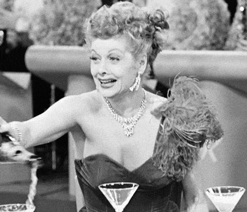 A woman in an elegant dress pours liquid from a bottle into martini glasses, smiling widely. The setting is a classy, well-lit venue.