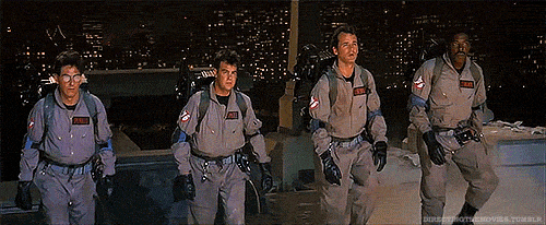 Four Ghostbusters in gray jumpsuits stand alert on a rooftop at night, with a city skyline glowing behind them.
