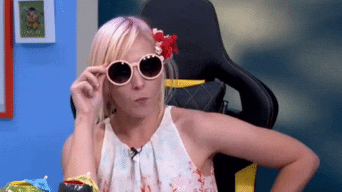 Woman with blonde hair and sunglasses, wearing a floral top, playfully adjusting glasses. Blue and white walls in the background.