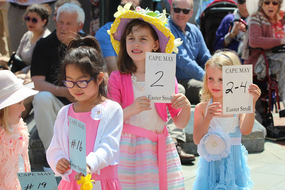 Children in colorful outfits hold "Cape May Easter Stroll" signs. Bright dresses, festive mood; crowd watches in a sunny outdoor setting.