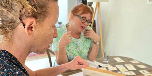 Deux femmes participent à une activité pédagogique avec des cartes illustrées disposées sur une table, pendant un cours de français en immersion chez Virginie in Provence