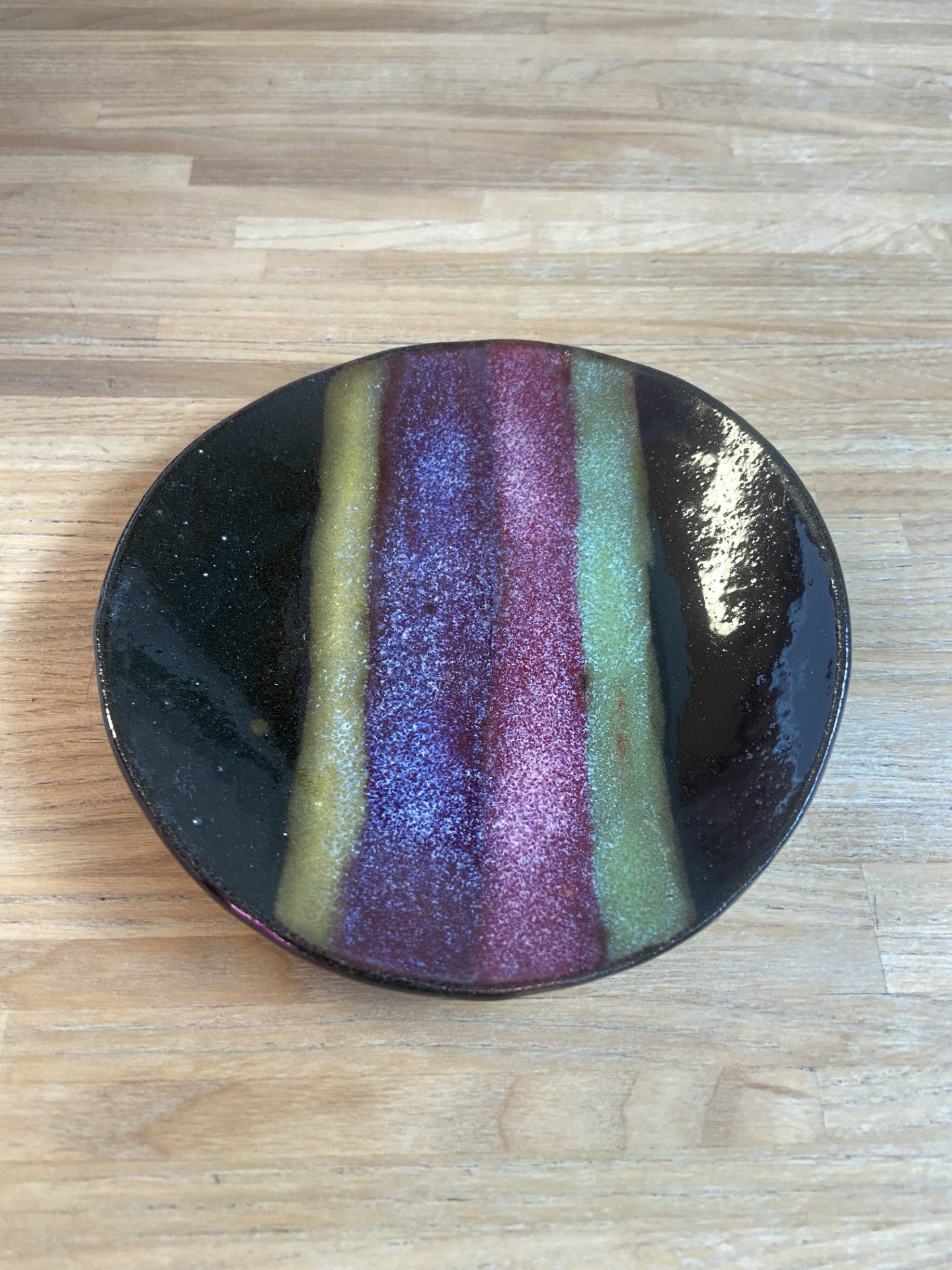 Handcrafted ceramic dish with deep plum and moss-green glaze stripes on a dark, glossy base, styled on natural wood.