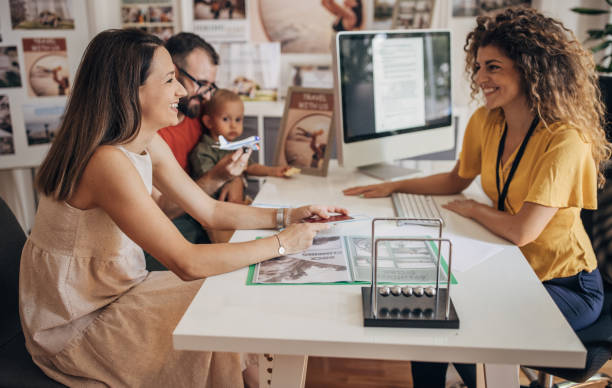 Smiling couple with baby consults woman at travel agency desk. Computer screen and travel brochures visible. Warm, inviting atmosphere.