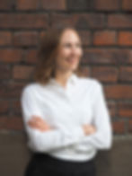 Folksin Counsel Lila Kallio seisoo valkoisessa paidassa hymyilee ja seisoo ruskeaa tiiliseinää vasten. Aurinkoinen ja rento tunnelma.