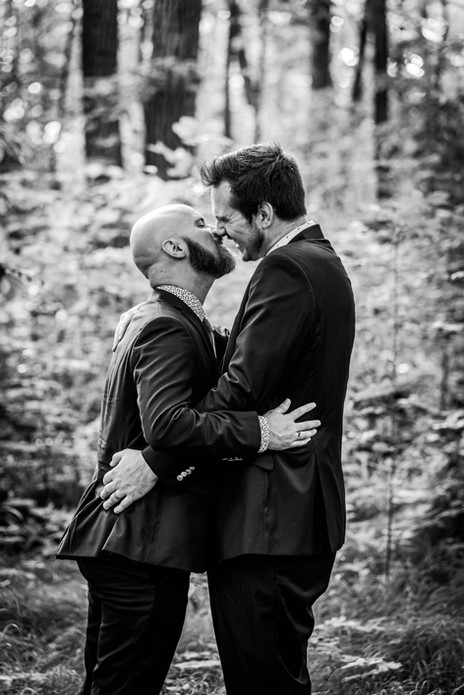 a black and white photo of two men in suits about to kiss. They are surround by trees in a soft, inviting looking forest.