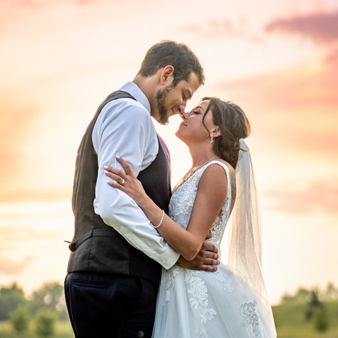 a bride and groom embrace and look at each other. Image shows waist up and a beautiful pink sunset in the background