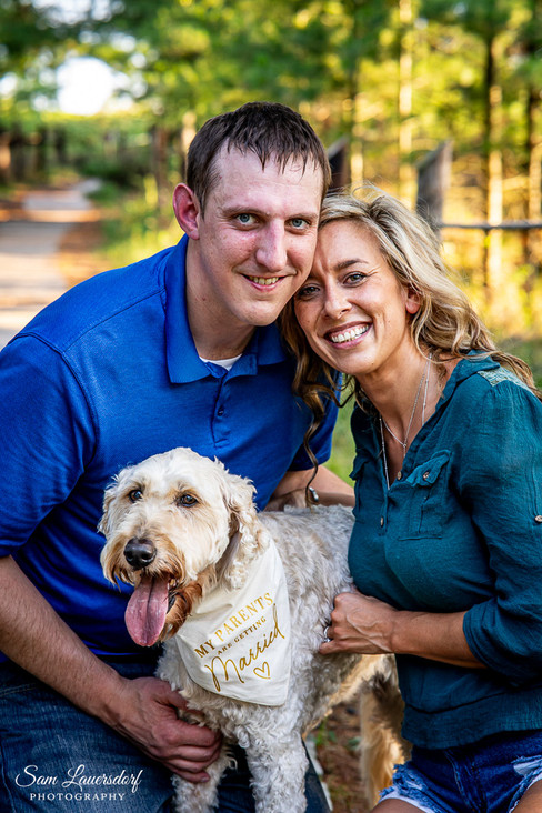 An engaged couple kneels down next to their dog who's wearing a "my parents are getting Married" bandana. Everyone is smiling at the camera.