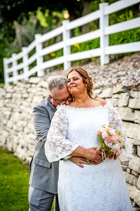 Groom in grey suit kisses wife in white long sleeve dress on the neck. A white picket fence and rock retaining wall are in the background