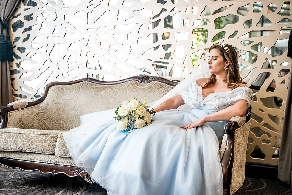 Bride in a light blue gown with a tiara, holding white roses, lounges on an ornate sofa. Intricate wall pattern and soft light in the background.