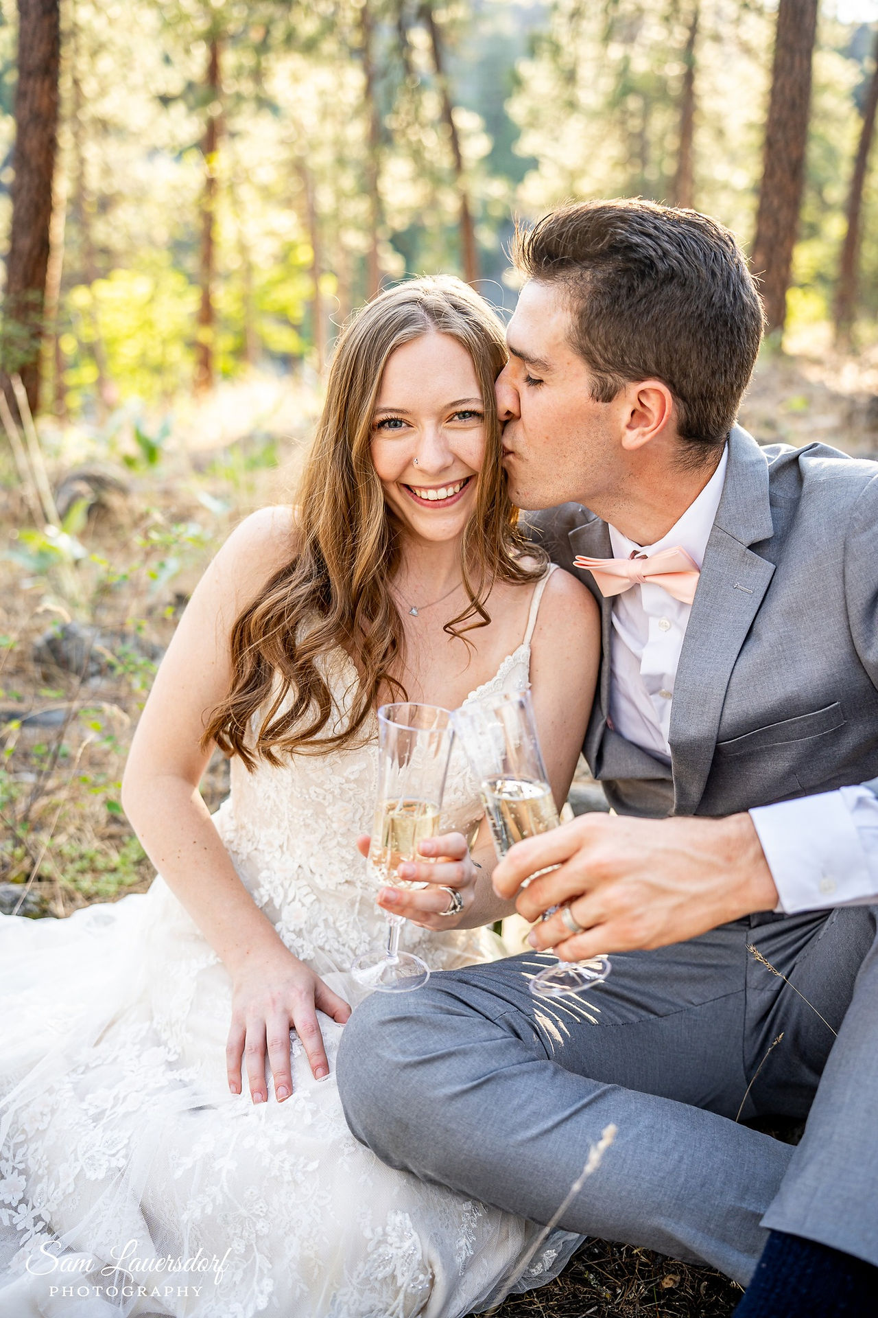 a couple sits close together on the group in the woods clinking glasses of champagne to celebrate their elopement