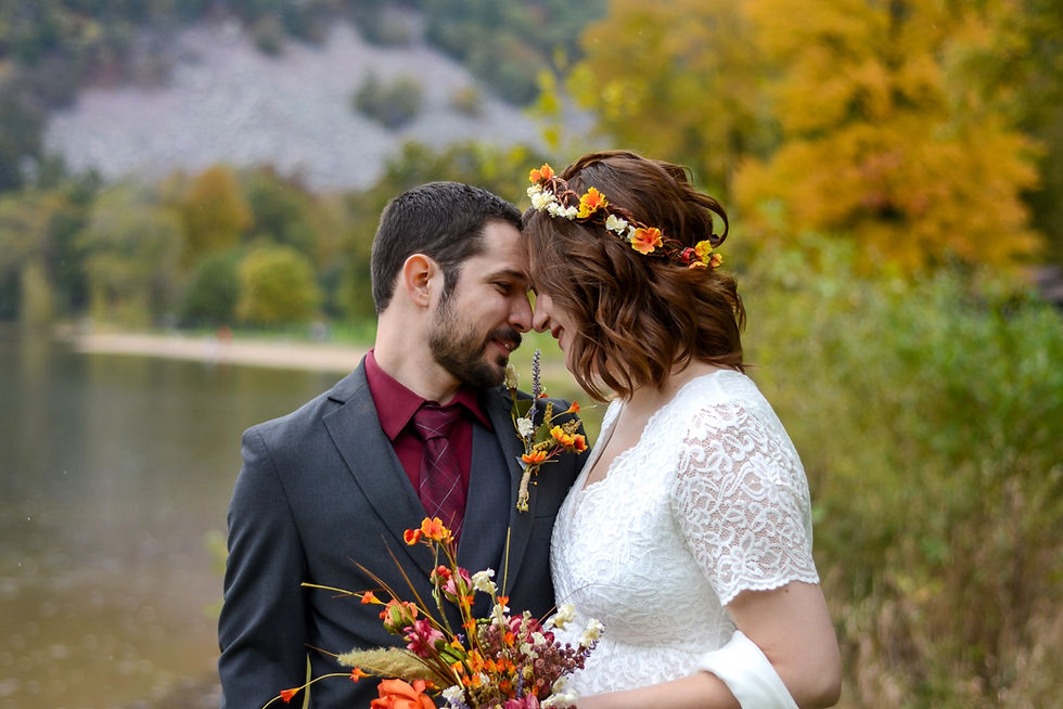 Bride and groom touch foreheads by a lakeside, holding a colorful bouquet, with autumn trees blurred in the background.