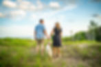 A couple stands, holding hands at Kohler Andrae State park looking at each other with Lake Michigan in the background. Their white poodle stands between them