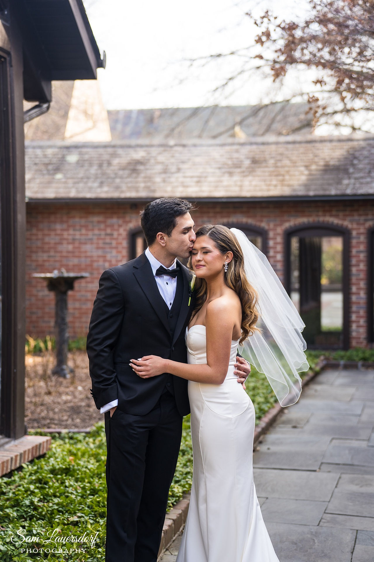 a groom in a black suit kisses bride on the forehead in the courtyard of Kohler American Club