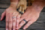 Two hands are laid flat on a piece of wood showing off wedding rings. A dogs paw is also laid by the human hands to show unity.