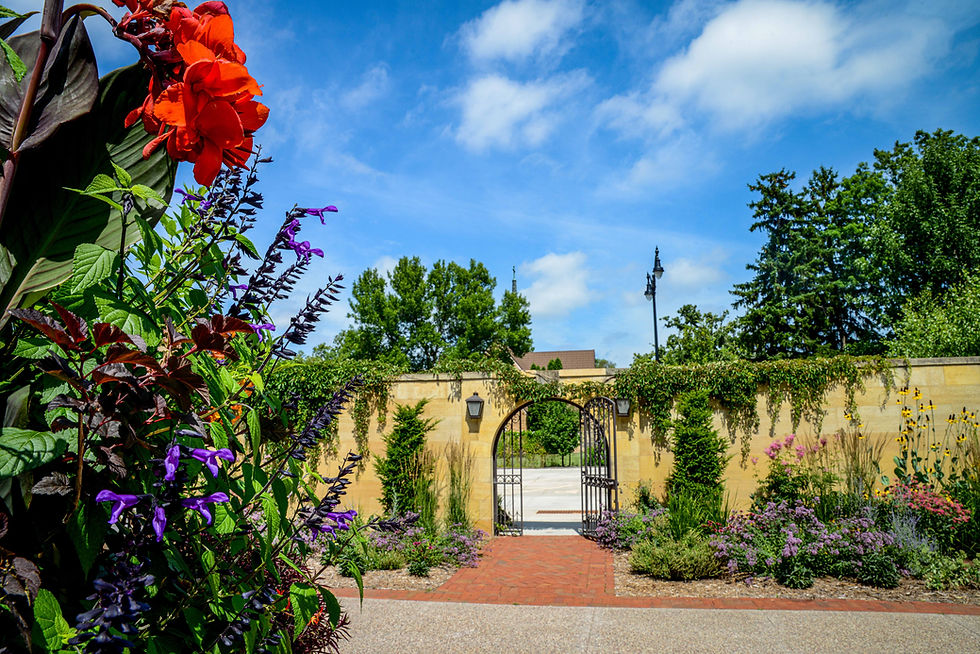 Vibrant garden with red and purple flowers in foreground. Walled archway leads to open area. Blue sky with clouds, lush greenery.