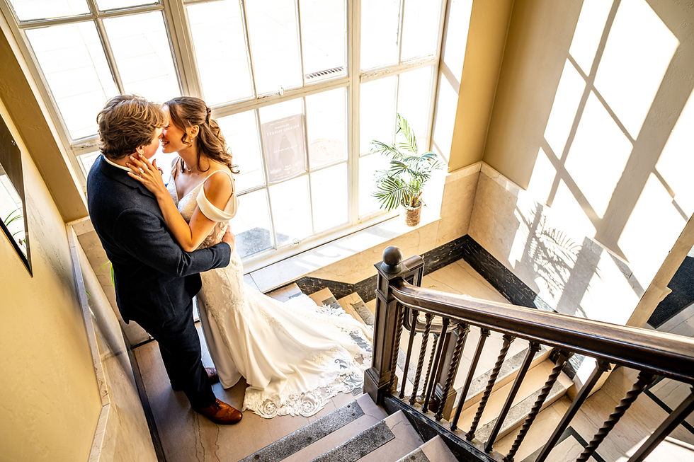 Couple kissing on a sunlit staircase in wedding attire, surrounded by elegant decor. A potted plant and window shadows add charm.