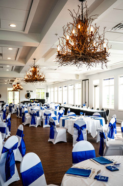Elegant event hall with round tables, white chairs adorned with blue sashes, and sparkling chandeliers.