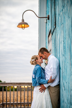 A bride and groom stand forehead to forehead under a light fixture against a light blue barn. The barn is Poplar Creek Barn in Oshkosh and features a cornfield in the background of the image