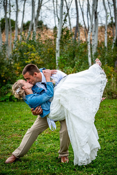 Groom dips and kisses his wife wearing a white dress and blue jean jacket. They are in front of a row of birch trees outside at Poplar Creek Barn